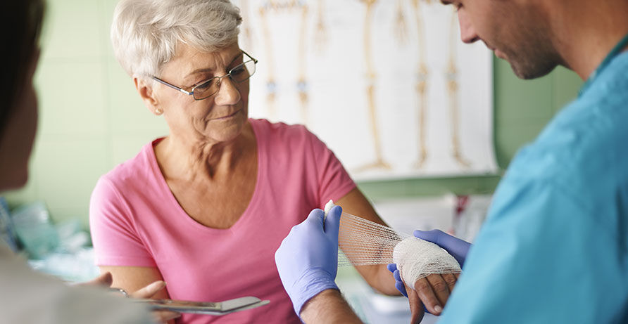 nitrilhandschuhe-krankenpflege Ein Krankenpfleger im blauen Einwegkittel trägt ebenfalls blaue Nitril Handschuhe. Er verbindet einer Seniorin die verletzte Hand.